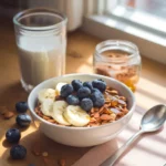 A bowl of granola topped with banana slices, fresh blueberries, and a drizzle of honey, placed near a glass of milk, a jar of cinnamon, and scattered almonds and blueberries on a wooden table by the window.