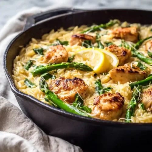 Creamy chicken orzo with asparagus and lemon slices in skillet on marble background