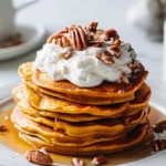Golden pumpkin pancakes stacked high with whipped cream and pecans on top